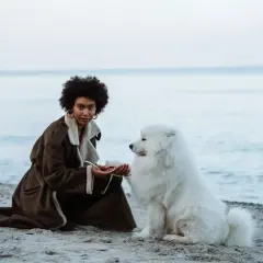 Kobieta z burzą włosów i biały pies na plaży dla psów. Spokojne morze w tle.