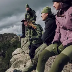 Grupa przyjaciół w zielonych i fioletowych kurtkach softshell i hardshell siedzi na skale, podziwiając górski krajobraz.