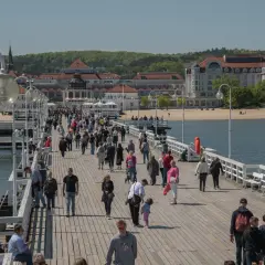 Tłum ludzi spaceruje po molo w Sopocie, jednym z najpopularniejszych gdańsk atrakcje. W tle widać plażę i zabytkowe budynki.