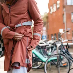Kobieta zapina swoją pomarańczową kurtkę przeciwdeszczową z odblaskowymi pasami. W tle stoją rowery.