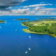 Wspaniałe wakacje nad wodą! Wiele żaglówek pływa po błękitnym jeziorze, otoczonym zielonymi wyspami i lasami.