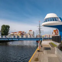 Niebieski most nad kanałem, przy którym znajduje się futurystyczna budowla i żaglowiec. Promenada nad morzem tętni życiem.