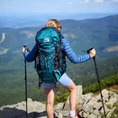 Kobieta z turkusowym plecakiem w góry, z kijkami trekkingowymi, podziwia panoramę.