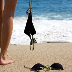 Nogi na piaszczystej plaży, obok czarny biustonosz. W tle błękitne morze i biała piana fal. Idealne miejsce na plażę nudystów.