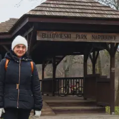 Kobieta w białej czapce i czarnej kurtce przed drewnianą wiatą z napisem "Białowieski Park Narodowy". To jedna z **hajnówka atrakcje**.