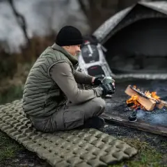Mężczyzna siedzi przy ognisku na dmuchanym materacu, zastanawiając się, jaki materac do namiotu będzie najlepszy na nocleg w terenie.
