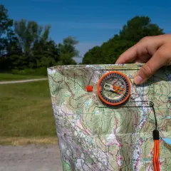 Ręka trzymająca mapę i kompas, kluczowe dla orientacji w terenie. W tle zielony krajobraz i błękitne niebo.