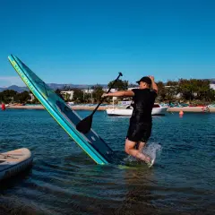 Mężczyzna w wodzie próbuje utrzymać równowagę na desce sup, która przechyla się na bok. W tle plaża i inne deski sup.