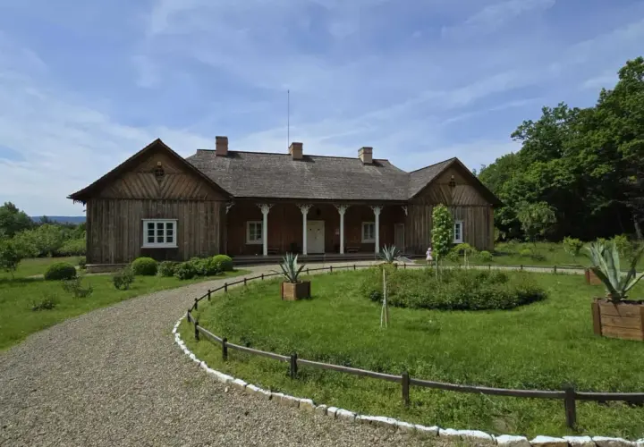 Drewniany dworek w skansenie Sanok, otoczony zielenią i kamienistą ścieżką, zaprasza do podróży w przeszłość.