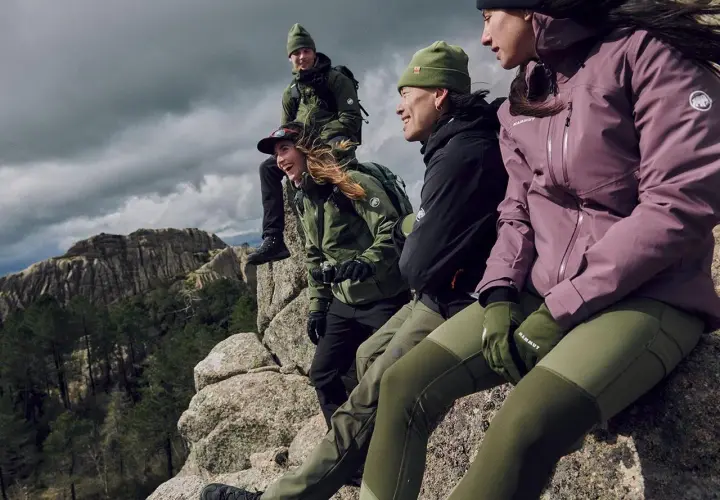 Grupa przyjaciół w zielonych i fioletowych kurtkach softshell i hardshell siedzi na skale, podziwiając górski krajobraz.