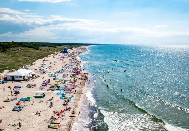 Plaża w Darłowie z turystami, parasolami i namiotami, w tle morze i las.