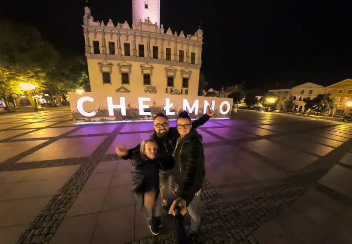 Rodzina pozuje do zdjęcia na tle napisu "Chełmno" i ratusza nocą.