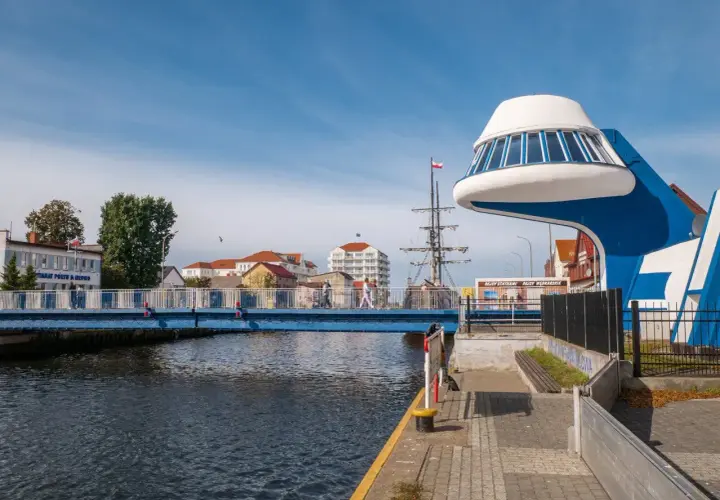 Niebieski most nad kanałem, przy którym znajduje się futurystyczna budowla i żaglowiec. Promenada nad morzem tętni życiem.