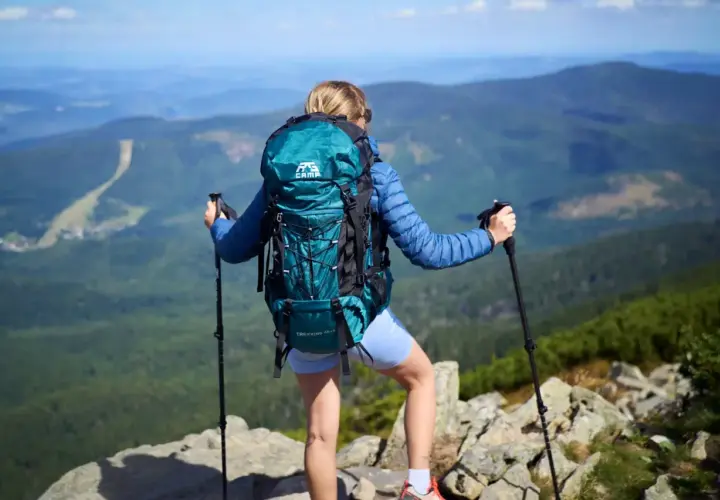 Kobieta z turkusowym plecakiem w góry, z kijkami trekkingowymi, podziwia panoramę.