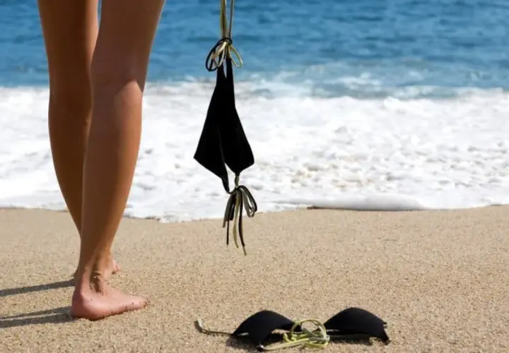 Nogi na piaszczystej plaży, obok czarny biustonosz. W tle błękitne morze i biała piana fal. Idealne miejsce na plażę nudystów.