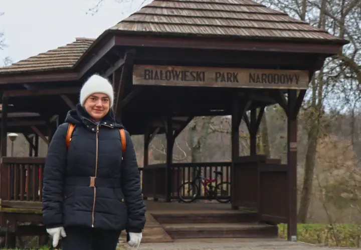 Kobieta w białej czapce i czarnej kurtce przed drewnianą wiatą z napisem "Białowieski Park Narodowy". To jedna z **hajnówka atrakcje**.