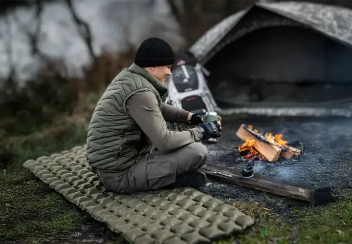 Mężczyzna siedzi przy ognisku na dmuchanym materacu, zastanawiając się, jaki materac do namiotu będzie najlepszy na nocleg w terenie.