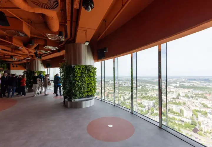 Wnętrze wieży widokowej we Wrocławiu z panoramicznym widokiem na miasto. Grupa ludzi zwiedza, podziwiając zieleń i architekturę.