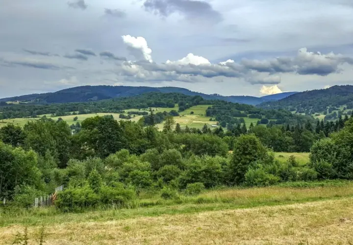 Zielone wzgórza i lasy w Węgierskiej Górce zapraszają na piesze wędrówki. Widok na dolinę i chmury na niebie.