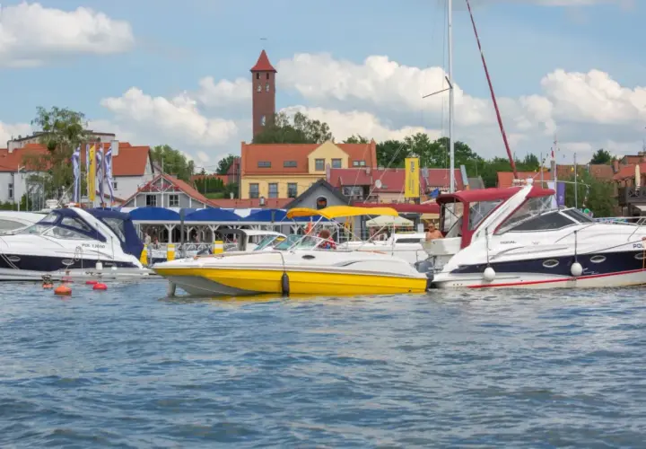 Żółta i białe łodzie motorowe cumują w porcie, w tle zabudowania i wieża. Idealne miejsce na bon Warmia Mazury.
