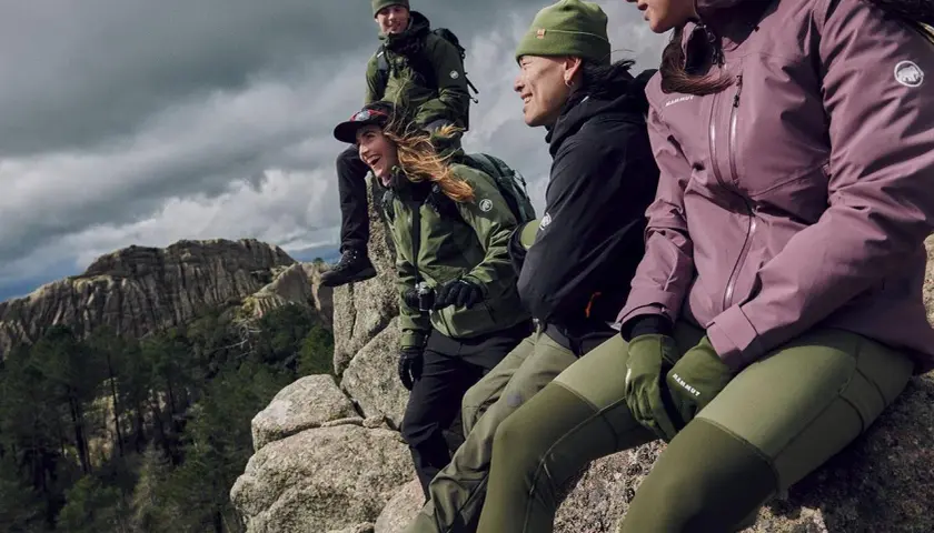 Grupa przyjaciół w zielonych i fioletowych kurtkach softshell i hardshell siedzi na skale, podziwiając górski krajobraz.