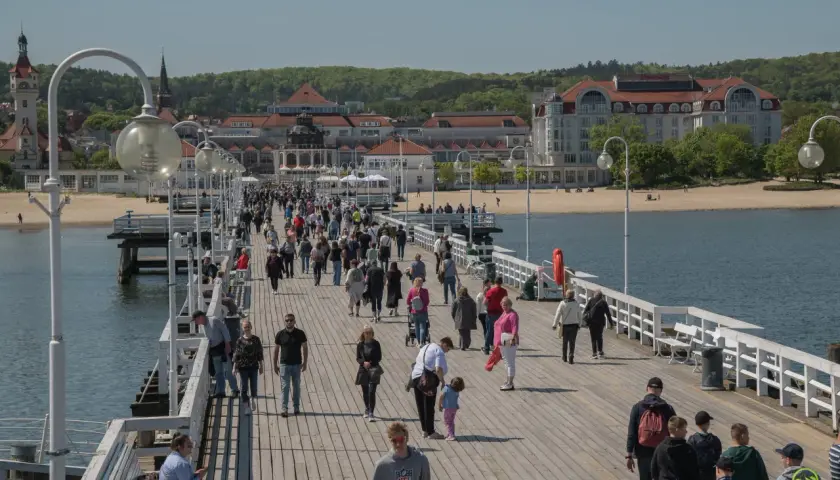 Tłum ludzi spaceruje po molo w Sopocie, jednym z najpopularniejszych gdańsk atrakcje. W tle widać plażę i zabytkowe budynki.
