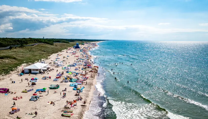 Plaża w Darłowie z turystami, parasolami i namiotami, w tle morze i las.