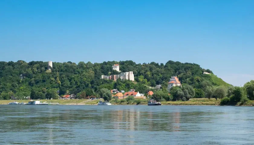 Widok na zamek i miasteczko na wzgórzu, z rzeką i łodziami na pierwszym planie. Kazimierz Dolny trasa zachwyca.