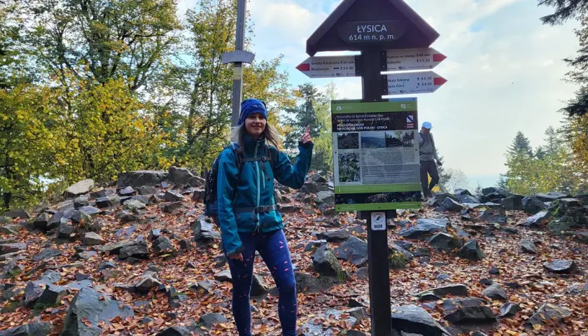Uśmiechnięta turystka na szczycie góry Łysica, obok drewnianego krzyża i tablicy informacyjnej. Jesienne liście pokrywają kamieniste podłoże.