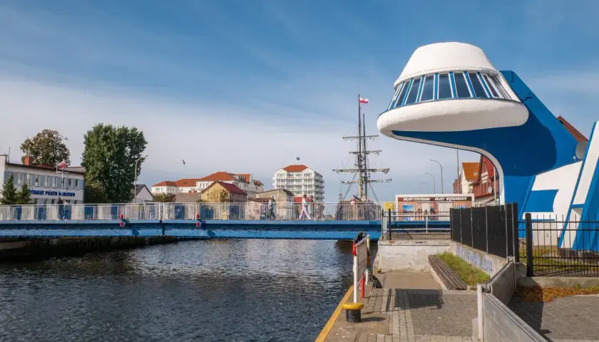 Niebieski most nad kanałem, przy którym znajduje się futurystyczna budowla i żaglowiec. Promenada nad morzem tętni życiem.