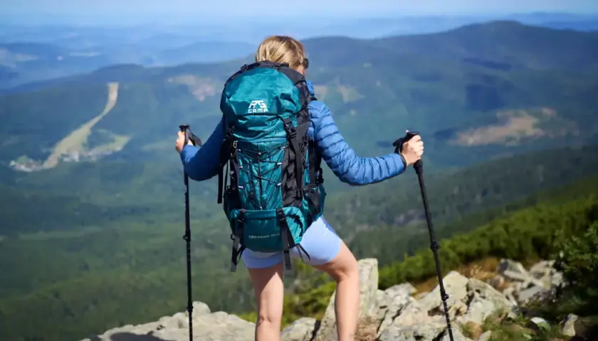 Kobieta z turkusowym plecakiem w góry, z kijkami trekkingowymi, podziwia panoramę.