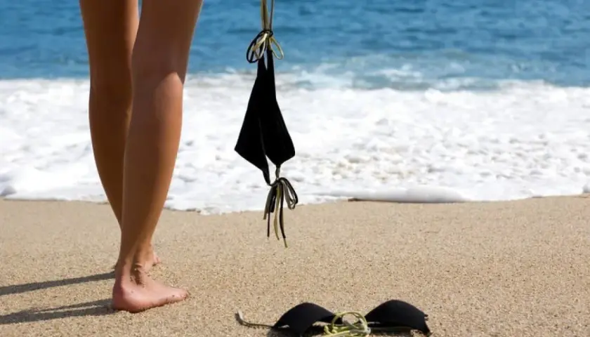 Nogi na piaszczystej plaży, obok czarny biustonosz. W tle błękitne morze i biała piana fal. Idealne miejsce na plażę nudystów.
