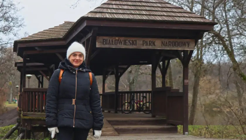 Kobieta w białej czapce i czarnej kurtce przed drewnianą wiatą z napisem "Białowieski Park Narodowy". To jedna z **hajnówka atrakcje**.