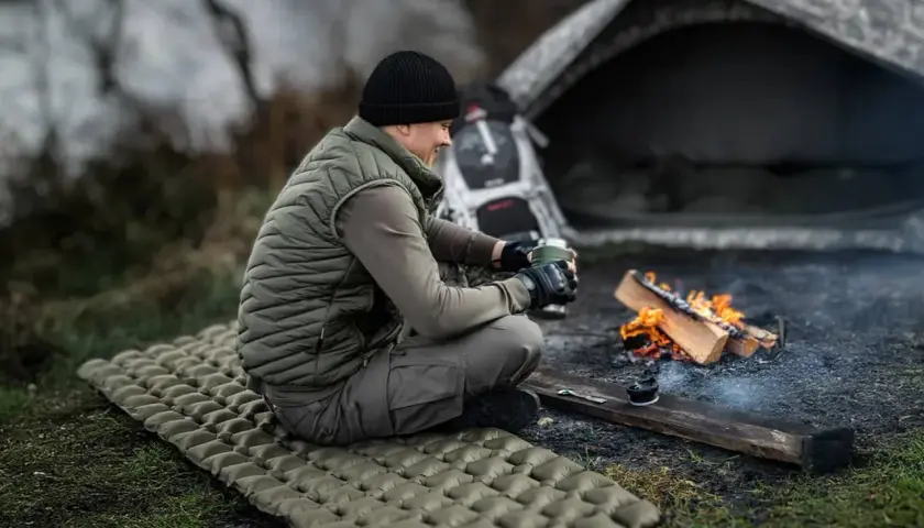 Mężczyzna siedzi przy ognisku na dmuchanym materacu, zastanawiając się, jaki materac do namiotu będzie najlepszy na nocleg w terenie.