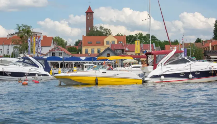 Żółta i białe łodzie motorowe cumują w porcie, w tle zabudowania i wieża. Idealne miejsce na bon Warmia Mazury.