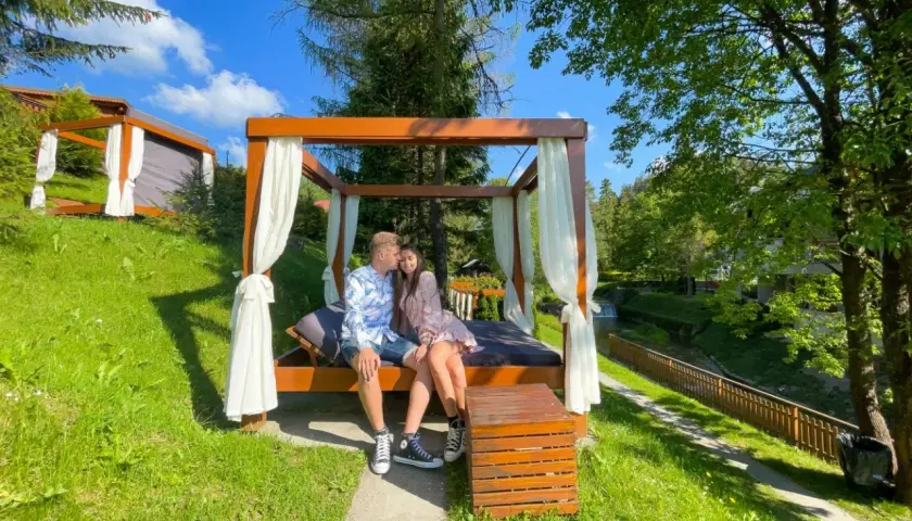 Para spędza romantyczny weekend w górach, siedząc na łóżku pod baldachimem, z widokiem na strumień i las.