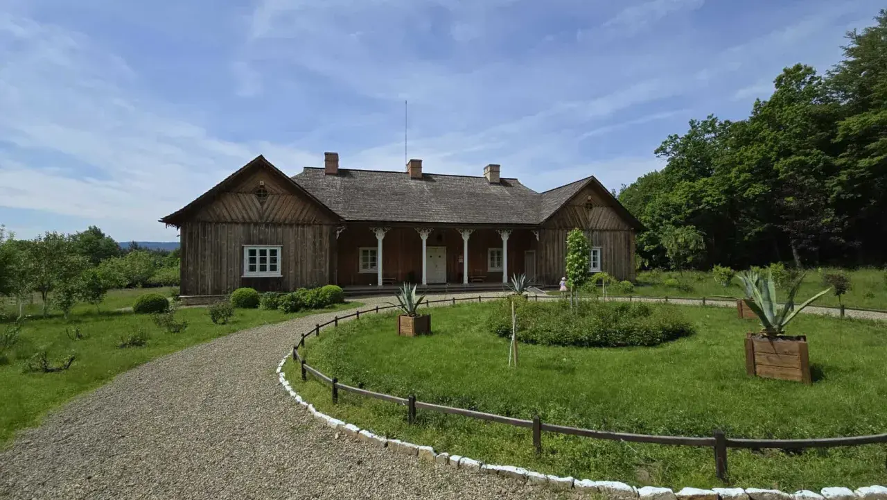 Drewniany dworek w skansenie Sanok, otoczony zielenią i kamienistą ścieżką, zaprasza do podróży w przeszłość.