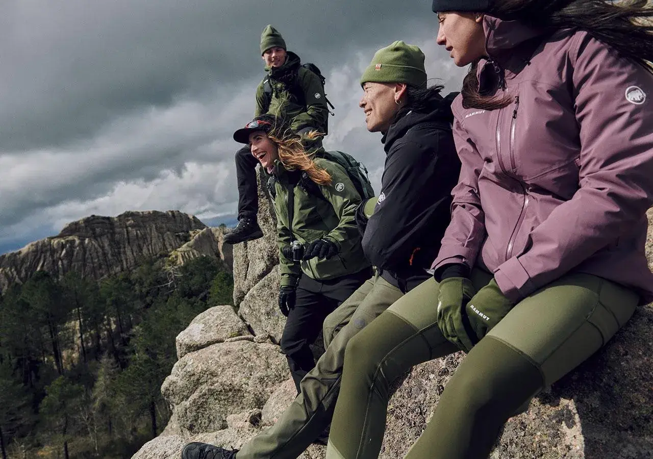 Grupa przyjaciół w zielonych i fioletowych kurtkach softshell i hardshell siedzi na skale, podziwiając górski krajobraz.