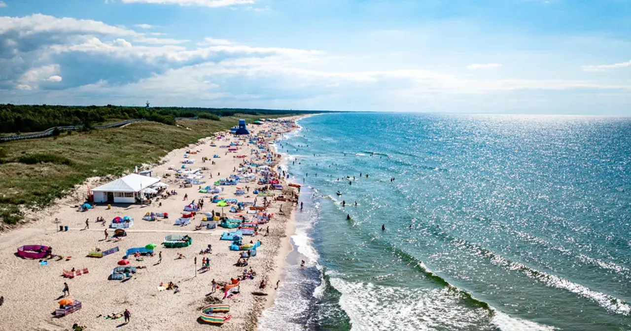 Plaża w Darłowie z turystami, parasolami i namiotami, w tle morze i las.