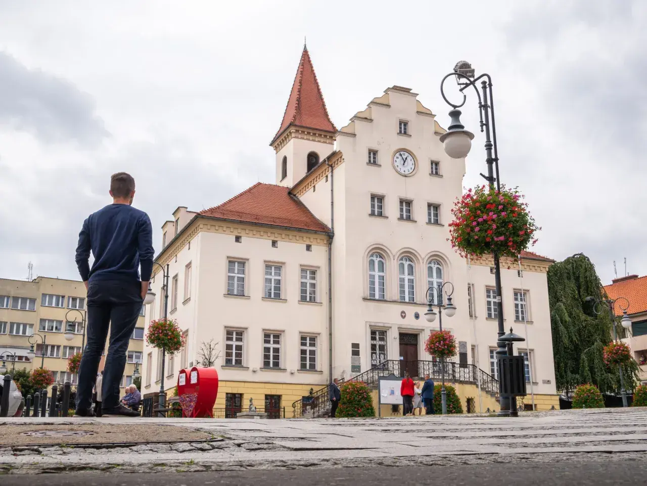 Mężczyzna podziwia ratusz w Trzebnicy, jedną z głównych atrakcji miasta. W tle czerwone serce i kwitnące kwiaty.