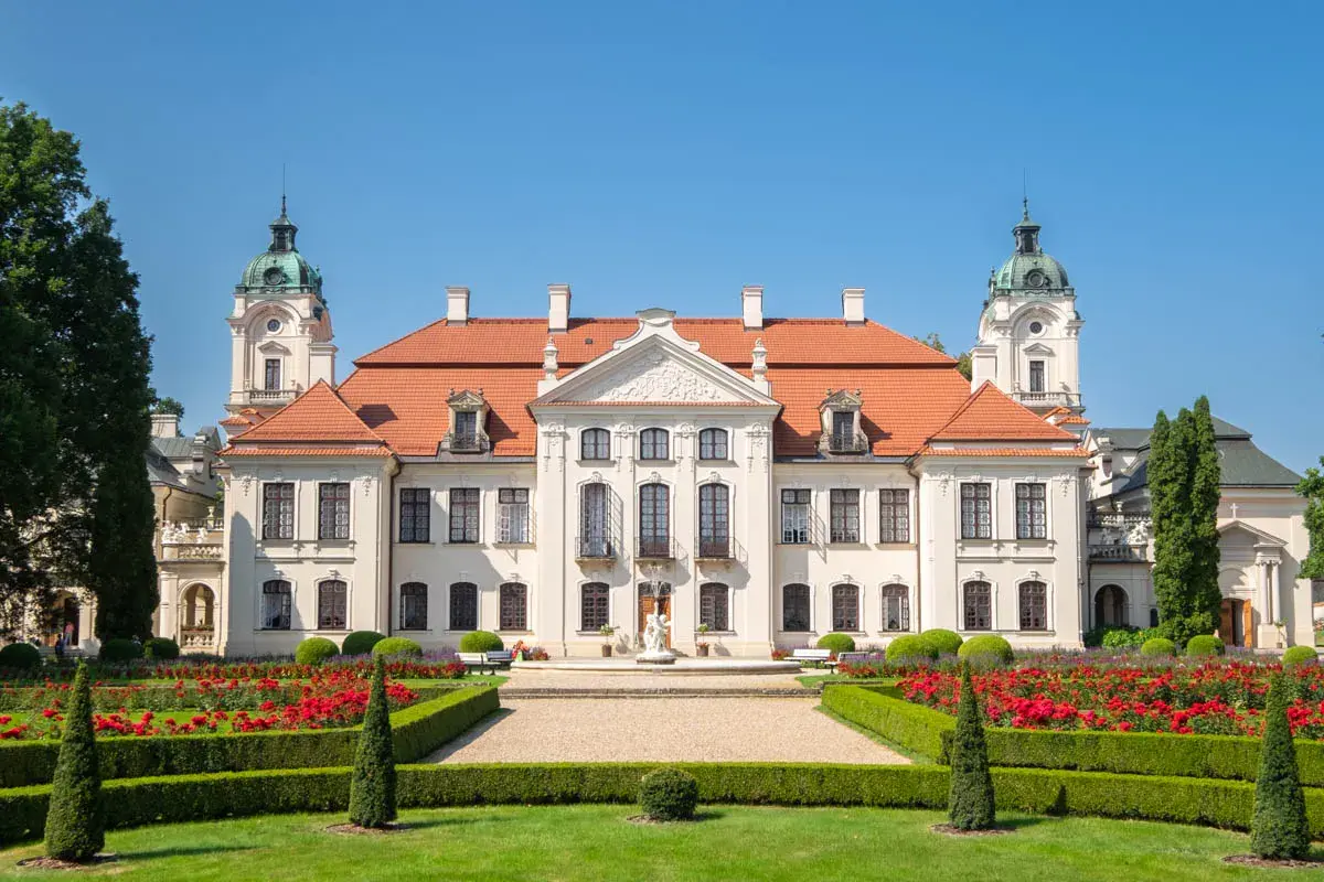Pałac Zamoyskich w Kozłówce, z czerwonym dachem i wieżyczkami, otoczony kwitnącym ogrodem z czerwonymi różami.
