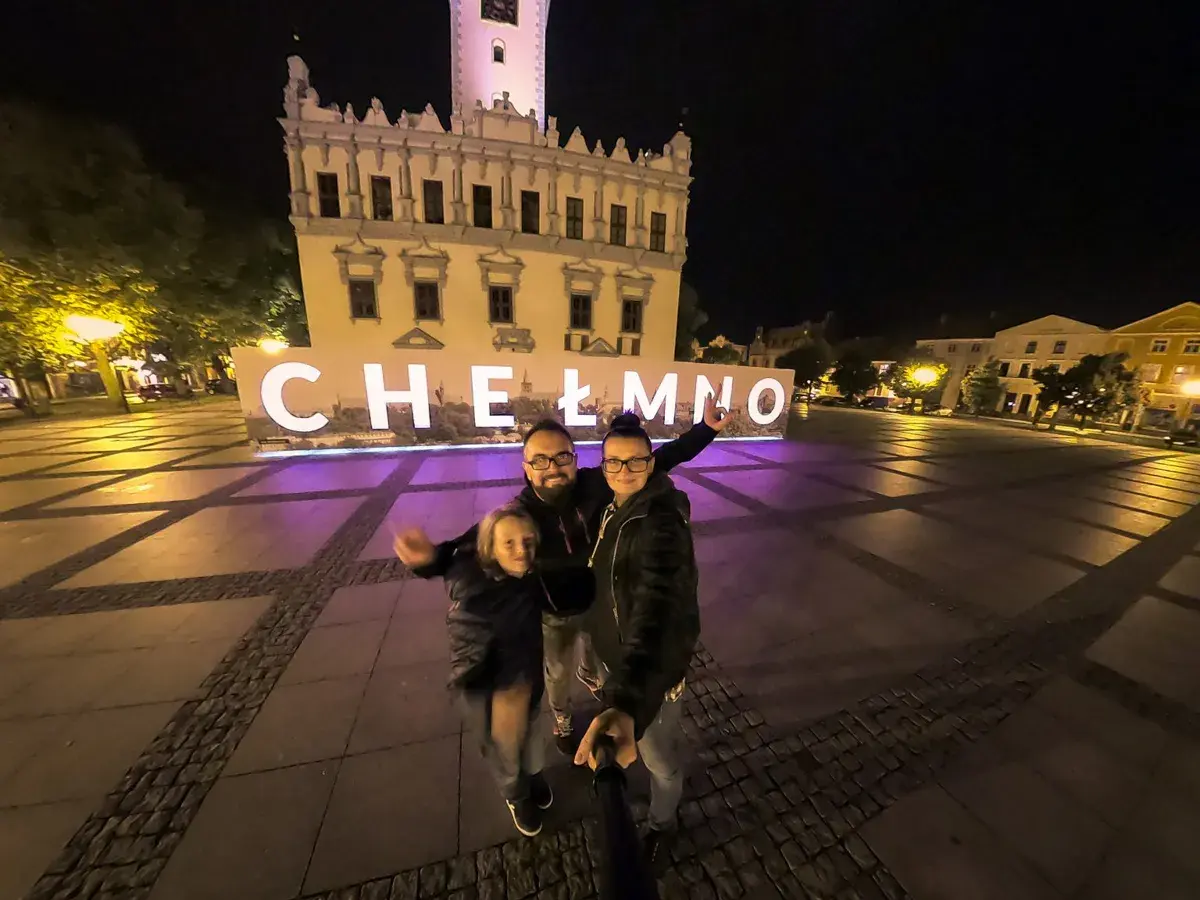 Rodzina pozuje do zdjęcia na tle napisu "Chełmno" i ratusza nocą.