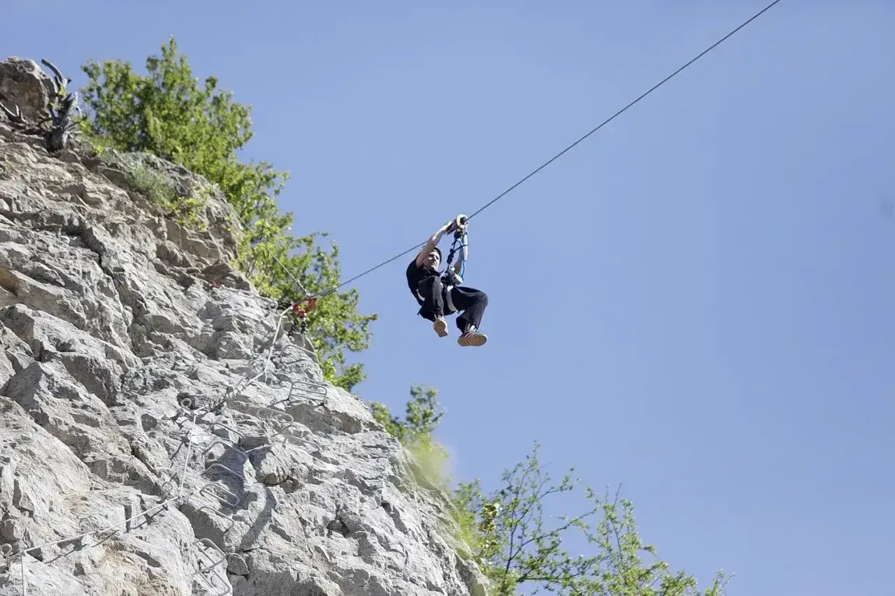 Człowiek zjeżdża na tyrolce obok skalnej ściany z ubezpieczeniami, jak na via ferrata polska.