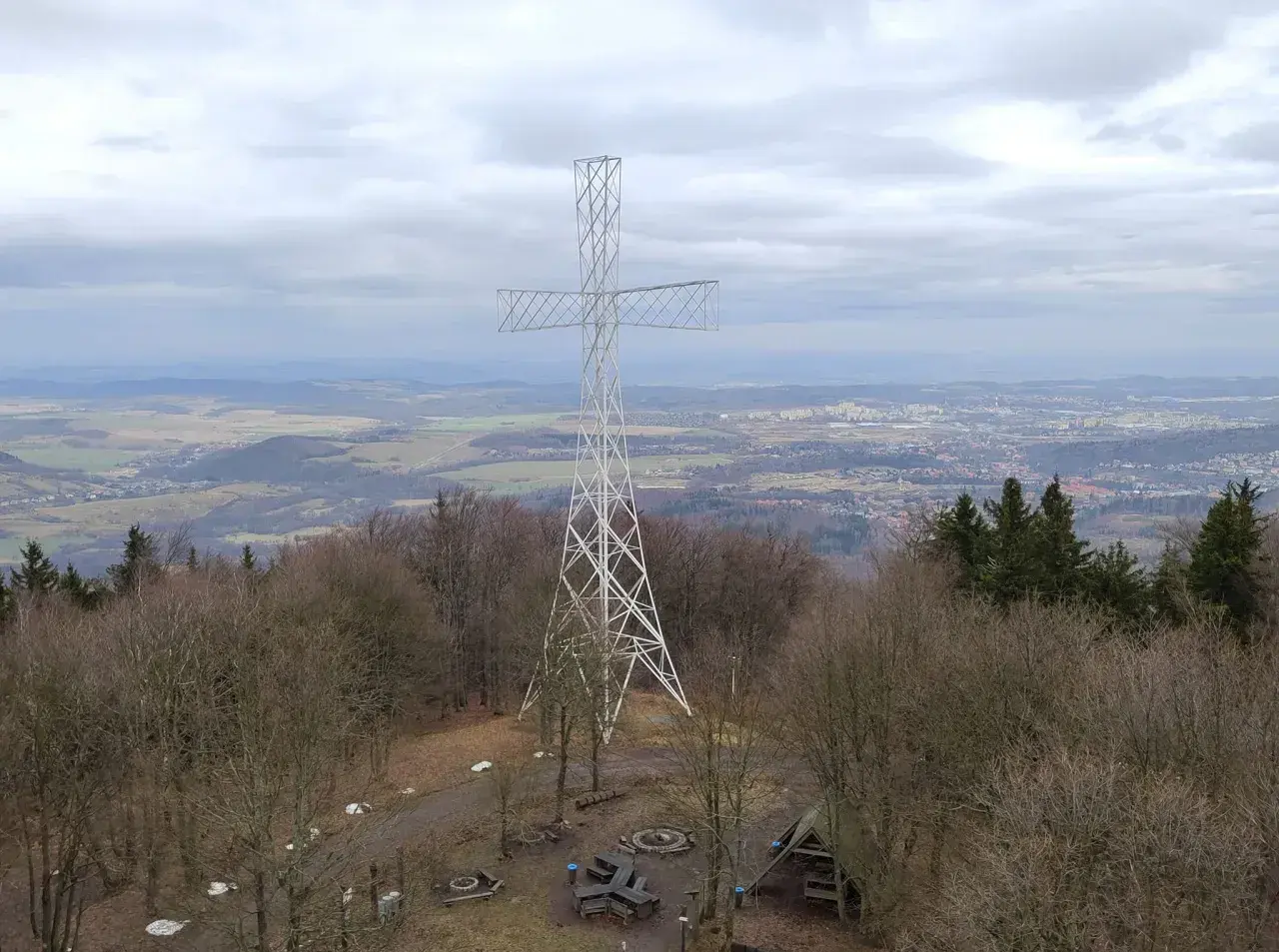 Wielki metalowy krzyż góruje nad lasem na **chełmiec szczyt**. W oddali rozciąga się panorama miasta i pagórkowatego krajobrazu.