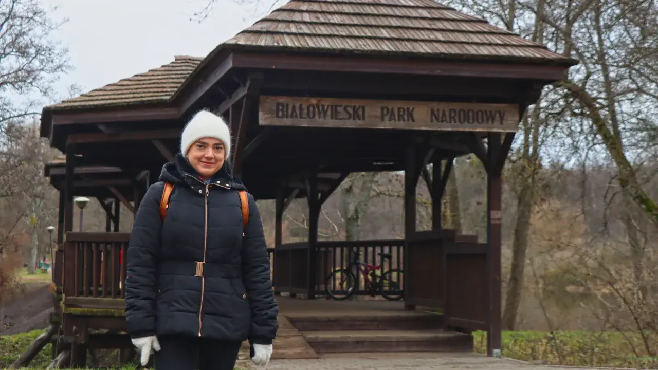Kobieta w białej czapce i czarnej kurtce przed drewnianą wiatą z napisem "Białowieski Park Narodowy". To jedna z **hajnówka atrakcje**.