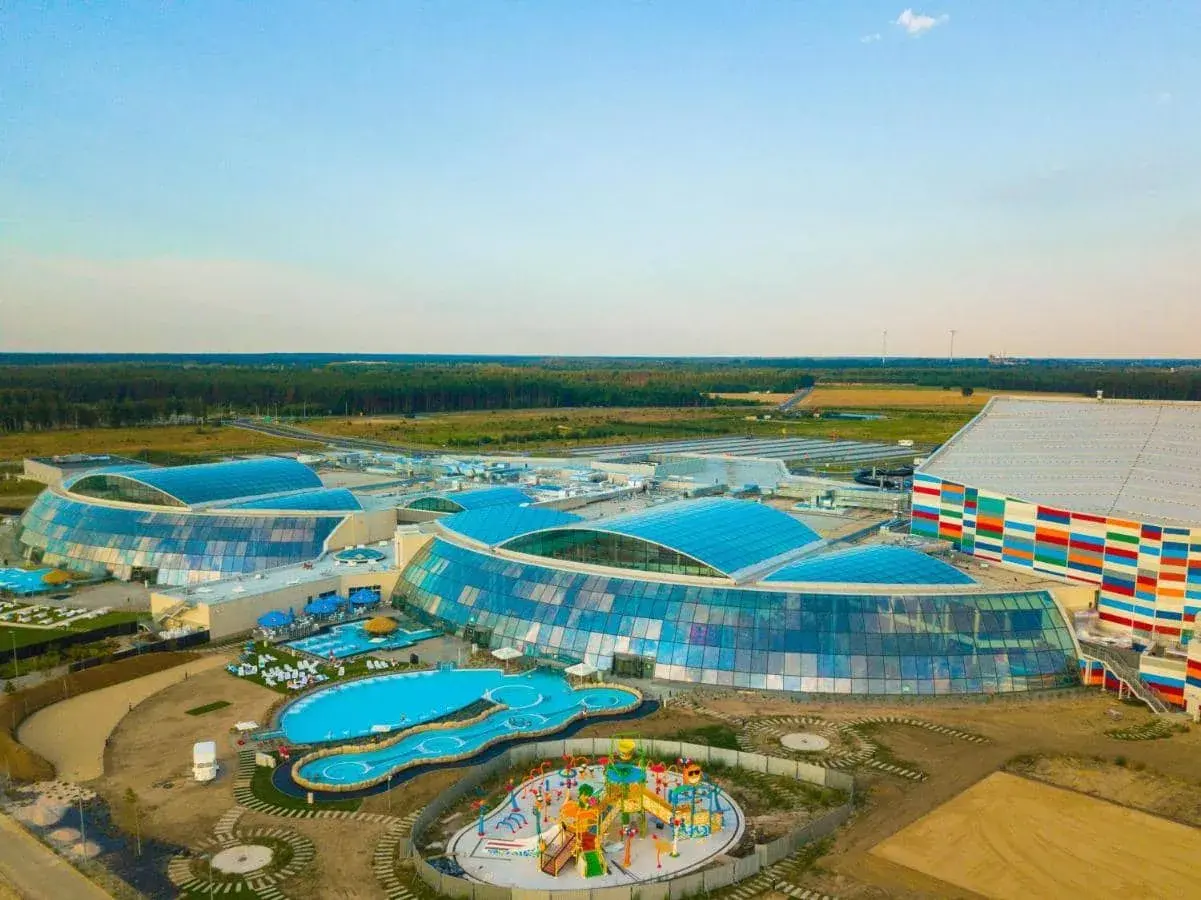 Największy aquapark w Polsce z błękitnymi, szklanymi dachami i kolorową fasadą, otoczony zielenią i basenami.