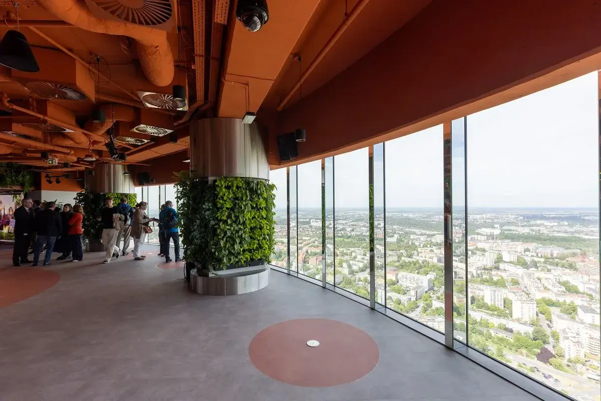Wnętrze wieży widokowej we Wrocławiu z panoramicznym widokiem na miasto. Grupa ludzi zwiedza, podziwiając zieleń i architekturę.
