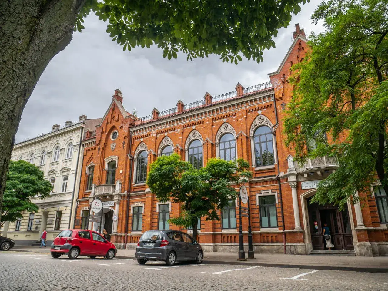 Kamienice w Radomiu, w tym Bank Polski, to ciekawe **radom atrakcje**. Dwa samochody zaparkowane na brukowanej ulicy.