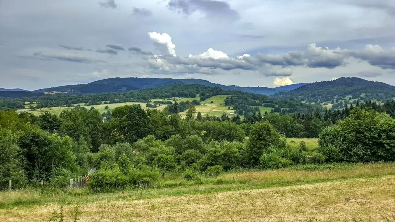 Zielone wzgórza i lasy w Węgierskiej Górce zapraszają na piesze wędrówki. Widok na dolinę i chmury na niebie.