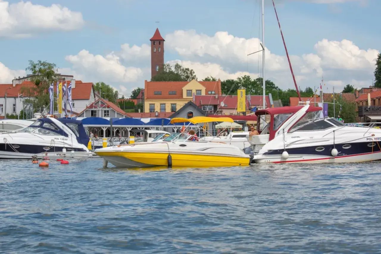 Żółta i białe łodzie motorowe cumują w porcie, w tle zabudowania i wieża. Idealne miejsce na bon Warmia Mazury.
